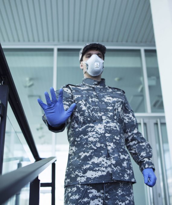Soldier in military uniform with rubber gloves and face protection mask guarding hospital doors and gesturing stop sign.
